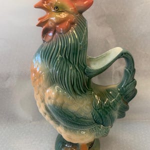 May include: A ceramic rooster pitcher with a green, yellow, and orange glaze. The rooster has a red comb and wattle.