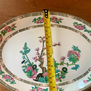May include: White oval serving platter with a green and pink floral design. The platter has a gold trim and a repeating pattern of green and gold squares around the edge. A measuring tape is placed next to the platter to show its size.