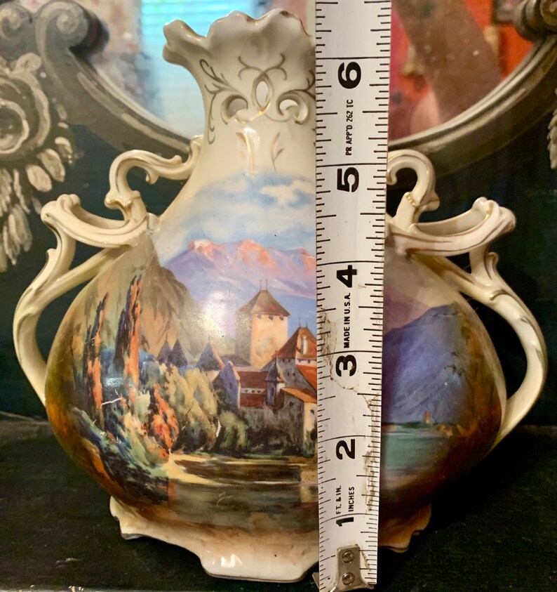 May include: A white porcelain vase with a hand-painted scene of a castle and mountains. The vase has two handles and is decorated with gold accents. It measures approximately 5 inches tall.