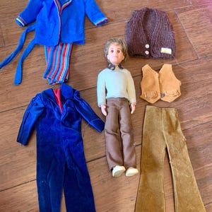 May include: A doll with brown hair and a blue shirt, wearing brown pants and white shoes. The doll is surrounded by various clothing items, including a blue jacket, a brown sweater vest, a brown corduroy vest, and a pair of brown pants.