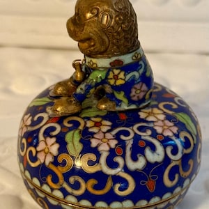 May include: A small, round, blue cloisonné box with a brass monkey figure on top. The box is decorated with floral and scroll patterns in white, pink, yellow, and green. The monkey is wearing a blue and green jacket.