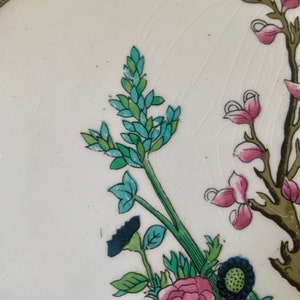 May include: Close-up of a floral design on a white ceramic plate. The design features a blue and green flower with a pink flower in the background.