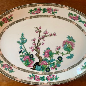 May include: White oval serving platter with a green, pink, and black floral design. The design features a central tree with branches and leaves, surrounded by smaller floral motifs. The border of the platter is decorated with a repeating pattern of green and gold.