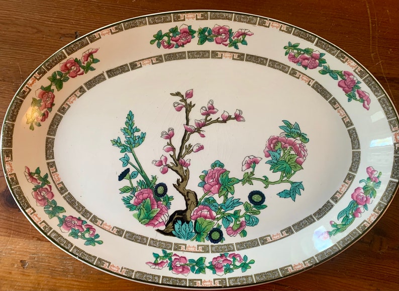 May include: White oval serving platter with a green, pink, and black floral design. The design features a central tree with branches and leaves, surrounded by smaller floral motifs. The border of the platter is decorated with a repeating pattern of green and gold.