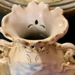 May include: A white porcelain vase with gold trim and intricate detailing. The vase has a scalloped rim and a delicate floral design.