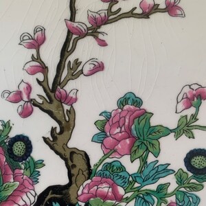 May include: A close-up of a white ceramic plate with a floral design. The design features pink and green flowers, a brown tree branch, and blue accents.