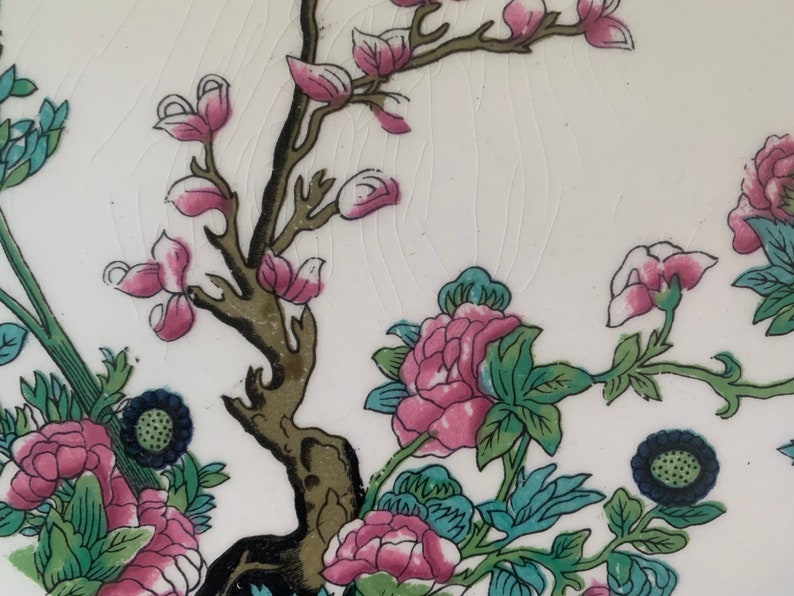 May include: A close-up of a white ceramic plate with a floral design. The design features pink and green flowers, a brown tree branch, and blue accents.
