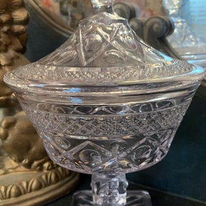 May include: A clear glass candy dish with a square base and a pineapple-shaped finial on the lid. The dish has a cut glass design.