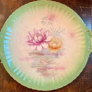 May include: A vintage green and white porcelain serving platter with floral designs. The platter has two handles and features a pink and yellow flower design with green leaves.