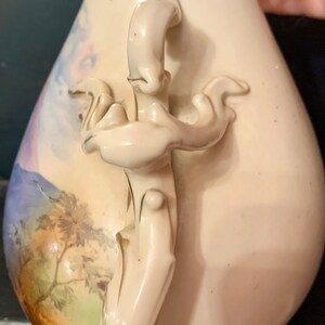May include: A white porcelain vase with a floral design and a detailed handle. The vase has a delicate, hand-painted landscape scene with a mountain range and trees.