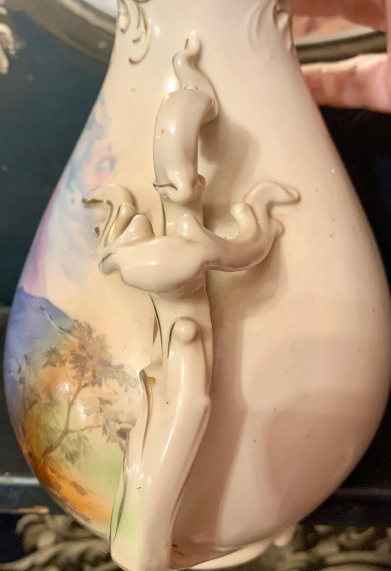 May include: A white porcelain vase with a floral design and a detailed handle. The vase has a delicate, hand-painted landscape scene with a mountain range and trees.