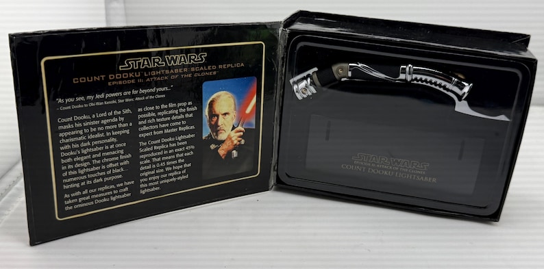 Puede incluir: Una r&eacute;plica del sable de luz Star Wars Count Dooku en una caja negra. El sable de luz tiene un dise&ntilde;o plateado y negro. La caja incluye texto y una imagen del Conde Dooku.