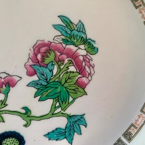 May include: A white ceramic plate with a floral design. The flowers are pink, green, and blue. The plate has a gold border with a repeating pattern.