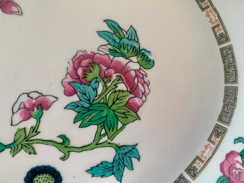 May include: A white ceramic plate with a floral design. The flowers are pink, green, and blue. The plate has a gold border with a repeating pattern.