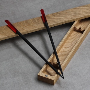 Może przedstawiać: Drewniane pudełko z otwartą pokrywą, ukazujące dwie pary czarnych pałeczek z czerwonymi końcówkami. Pałeczki leżą w pudełku.