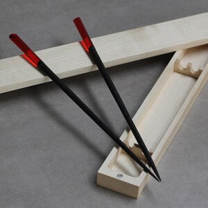 Może przedstawiać: Drewniane pudełko z pokrywką, zawierające dwie pary czarnych pałeczek z czerwonymi końcówkami. Pudełko wykonane jest z jasnego drewna i ma prosty design.