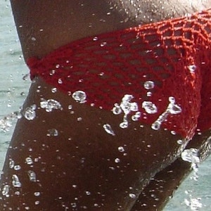 May include: A vibrant red bikini bottom featuring a distinct open-work crochet knit pattern. The garment is shown with water droplets splashing across the surface, highlighting the textured, knotted construction of the fabric and the side tie detail.