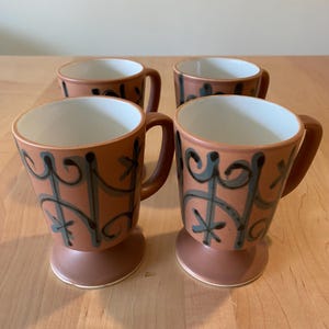 May include: Four ceramic mugs with a brown exterior and white interior. Each mug features a decorative design in gray and black. The mugs have a pedestal base and a curved handle. The mugs are displayed on a light brown wooden surface.