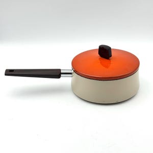 May include: A small, cream-colored saucepan with an orange lid and a dark brown handle. The lid has a matching dark brown knob. The handle is attached with a silver metal piece. The pan is on a white background.