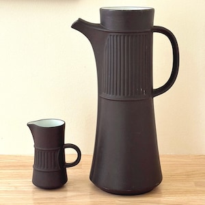 May include: A dark brown ceramic coffee carafe and creamer set. The carafe has a tall, slender body with a spout and handle. The creamer is smaller, with a similar design. Both have vertical ribbed detailing.