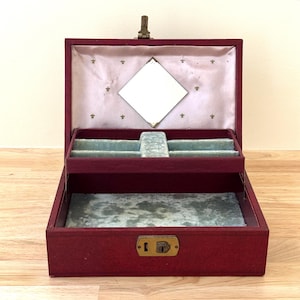 May include: Open burgundy jewelry box with a light blue interior and a small mirror. The box has a gold-colored clasp and keyhole. The interior is lined with light blue velvet and a light pink fabric with gold fleur-de-lis.