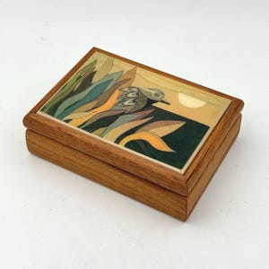 May include: A rectangular wooden box with a hinged lid featuring an inlaid graphic design of a bird among stylized leaves in shades of green, orange, and brown against a pale yellow sky with a circular sun. The box has a natural wood grain finish.