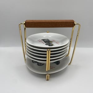 May include: A set of six white ceramic saucers with a vintage car design and red text. The saucers are stacked in a gold metal stand with a wooden handle.