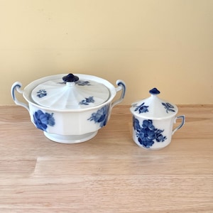 May include: A white porcelain sugar bowl and creamer set with blue floral designs. The sugar bowl has a lid and handles, while the creamer has a handle and a lid. Both pieces have a dark blue finial.