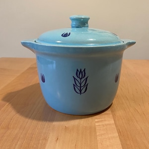 May include: A light blue ceramic casserole dish with a lid. The dish features dark blue tulip designs and handles on either side. The lid has a small knob and tulip designs. The casserole dish is sitting on a wooden surface.