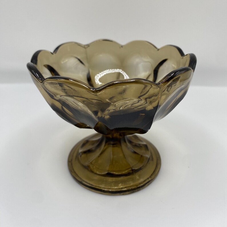 May include: A vintage brown glass pedestal bowl with a scalloped rim. The bowl has a round base and a stem. The glass has a smoky, translucent appearance, with darker areas adding depth. A decorative piece for serving or display.