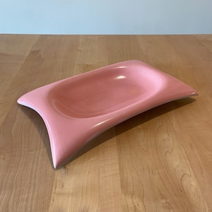 May include: A pink, rectangular ceramic dish with curved edges. The dish has a shallow, rounded interior and a glossy finish. The overall shape is elongated, with flared ends. The dish is likely for serving or display.