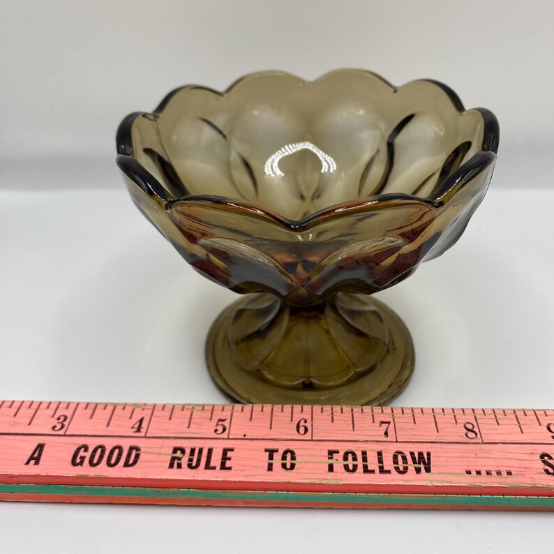 May include: An olive-green glass dessert dish with a scalloped rim and pedestal base. The dish has a flower-like design and is approximately 10 cm in diameter. The text "A GOOD RULE TO FOLLOW" is visible.