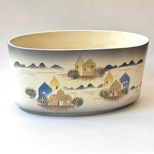 May include: An oval ceramic planter with a gray and white background and a painted scene of houses in a landscape. The houses are painted in shades of yellow, blue, and brown.