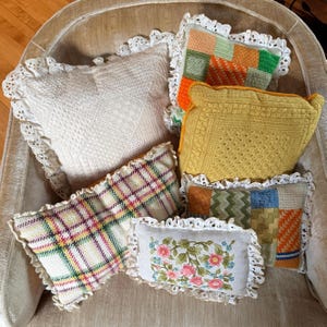 May include: A collection of decorative pillows with various designs. Pillows include a white, textured square pillow, a yellow crocheted pillow, and a plaid pillow. Each pillow is trimmed with a white lace border.