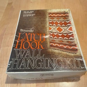 May include: A WonderArt latch hook wall hanging kit. The box features a finished size of 18 x 27 inches. The design on the box shows a geometric pattern in shades of brown, orange, and cream. The text "LATCH HOOK WALL HANGING KIT" is prominently displayed.