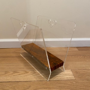 May include: A clear acrylic magazine rack with a wood shelf. The shelf is a dark brown with a marbled pattern. The rack has a modern design and is perfect for storing magazines, newspapers, or other reading materials.