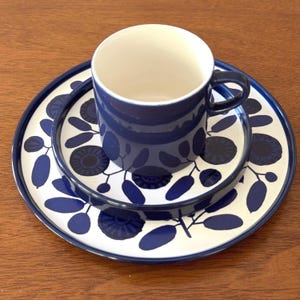 May include: A navy blue ceramic teacup with a handle sits on a matching saucer and a larger plate. The set features a white background with a repeating blue floral pattern. The cup and plates are on a wooden surface.