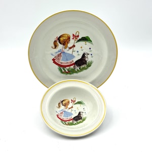 May include: A white ceramic plate and bowl set with a yellow rim. The plate and bowl feature a colorful illustration of a girl with a staff and a dog. The girl is wearing a blue dress and a red bow.