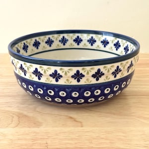 May include: A round ceramic bowl featuring a dark blue base with a repeating pattern of white circles with dark blue centers. The upper section displays a band of small blue four-petaled flowers alternating with light green leaf-like shapes.