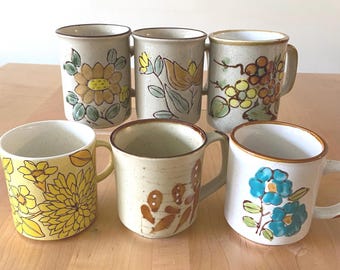 May include: A collection of six ceramic mugs with speckled off-white or yellow bases. Each mug features hand-painted floral designs in various colors including yellow, brown, and blue. The mugs have cylindrical bodies and side handles.