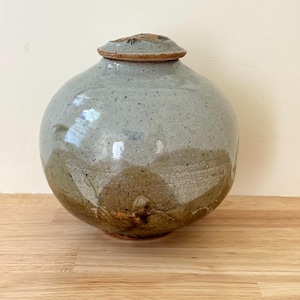 May include: A round ceramic jar with a lid, featuring a speckled light blue-grey glaze on the upper portion and a textured, earthy brown glaze on the lower section. The vessel has a smooth, bulbous body and a small, circular matching lid.