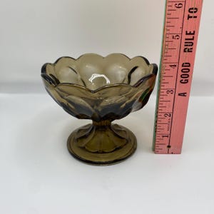 May include: A vintage, brown glass dessert dish with a scalloped rim and a pedestal base. The dish is approximately 7.6 cm tall. The glass has a slight amber tint, and the design is reminiscent of antique glassware.