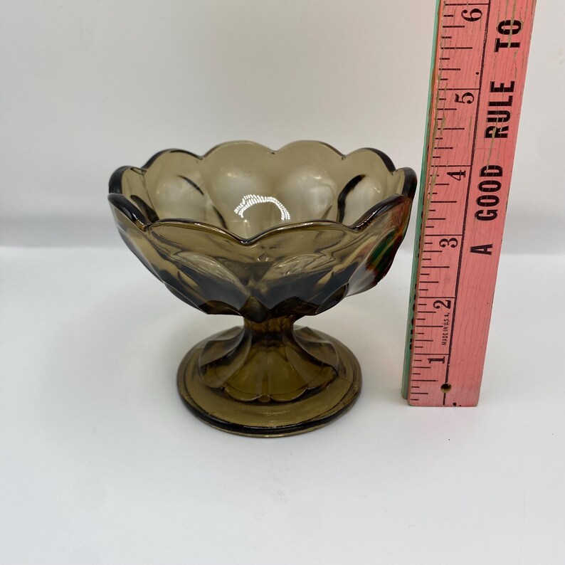 May include: A vintage, brown glass dessert dish with a scalloped rim and a pedestal base. The dish is approximately 7.6 cm tall. The glass has a slight amber tint, and the design is reminiscent of antique glassware.
