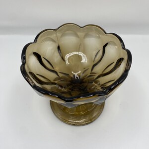 May include: A vintage, brown-tinted glass bowl with a scalloped rim and a pedestal base. The bowl has a floral design and is likely used for serving desserts or displaying decorative items. The glass has a glossy finish.