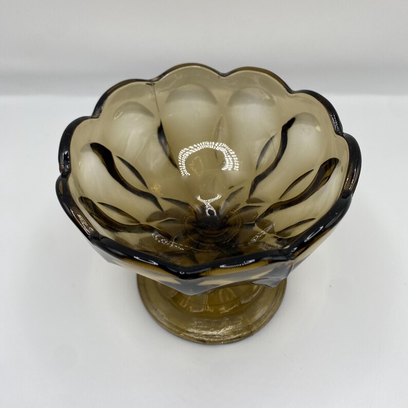 May include: A vintage, brown-tinted glass bowl with a scalloped rim and a pedestal base. The bowl has a floral design and is likely used for serving desserts or displaying decorative items. The glass has a glossy finish.