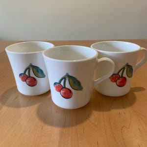 May include: Three white ceramic mugs with a cherry design. Each mug features a hand-painted illustration of red cherries with green leaves. The mugs have a simple, elegant design, perfect for coffee or tea.