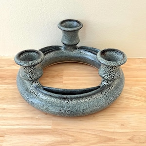 May include: A speckled gray ceramic candle holder with three circular candle holders. The candle holder is a ring shape with a small trough. The candle holders are cylindrical and have a speckled pattern.