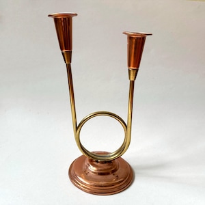 May include: A brass and copper candle holder with two candle cups. The candle holder has a circular base and two arms that curve upwards to hold the candle cups.