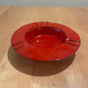 May include: A round, red ashtray with a glossy finish. The ashtray has a circular design with four grooves for resting cigarettes. The interior is a deep red, and the exterior has a slightly textured appearance.