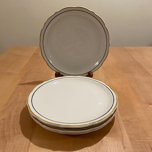 May include: A stack of three white plates with a scalloped edge and a gold rim. Each plate has a black line just inside the rim. One plate is displayed on a wooden stand.